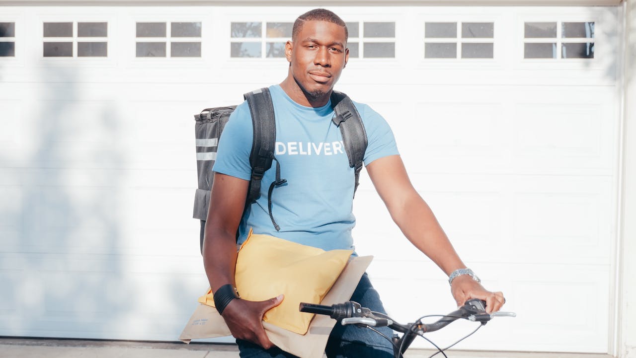 about-img African American courier on bicycle holding parcels, ready for delivery in a sunny outdoor setting.