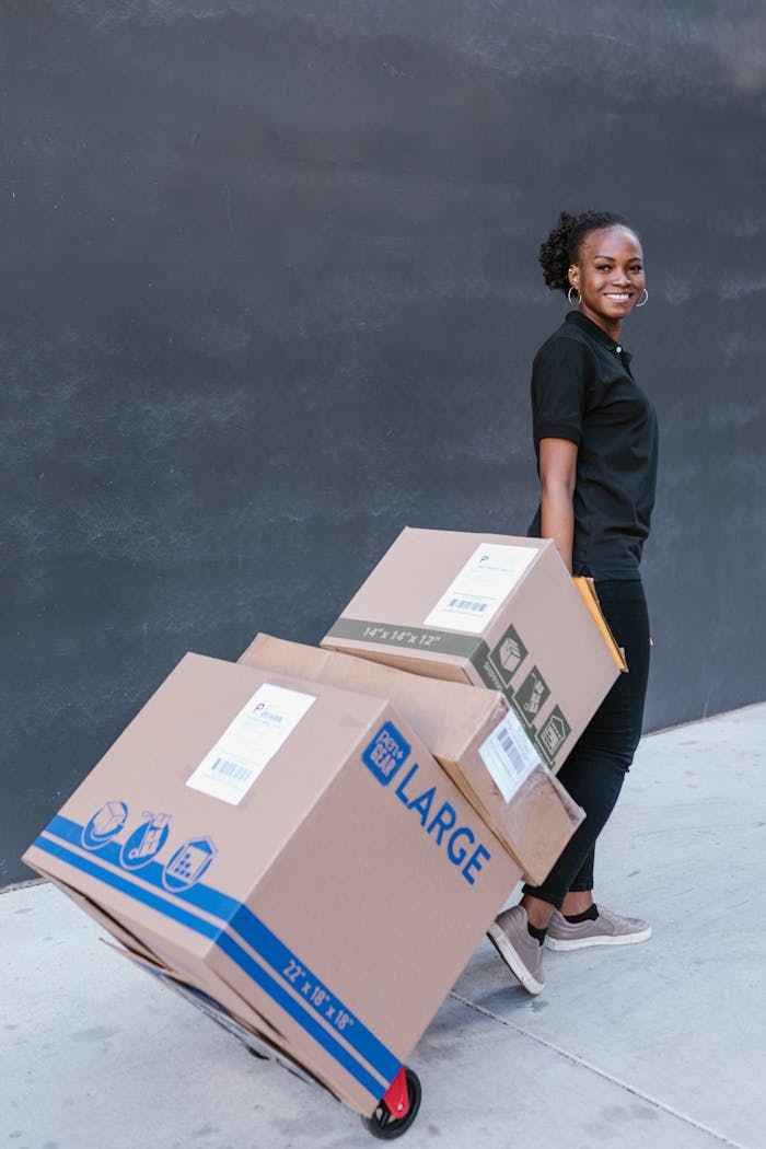 services-img Young woman delivering cardboard boxes on a dolly outdoors, smiling confidently.